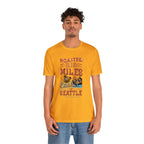Adult man modeling a golden yellow short-sleeve T-shirt printed with a colorful graphic that reads “Roasted 818 Miles East of Seattle,” featuring a steaming coffee cup and mountain lake illustration, shown against a plain white background.