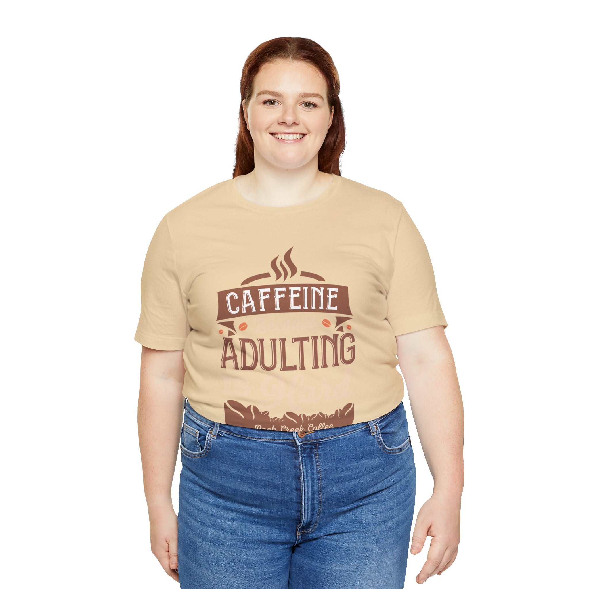 Adult woman smiling while modeling a soft cream short-sleeve T-shirt featuring the “Caffeine Because Adulting Is Hard” graphic in a stacked typographic design, shown against a plain white background.