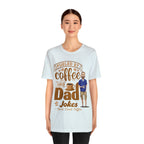 Adult woman modeling a light blue heather short-sleeve T-shirt with a coffee-themed printed graphic reading “Fueled by Coffee and Dad Jokes,” featuring a steaming coffee cup and cartoon dad illustration on the front, shown against a plain white background.