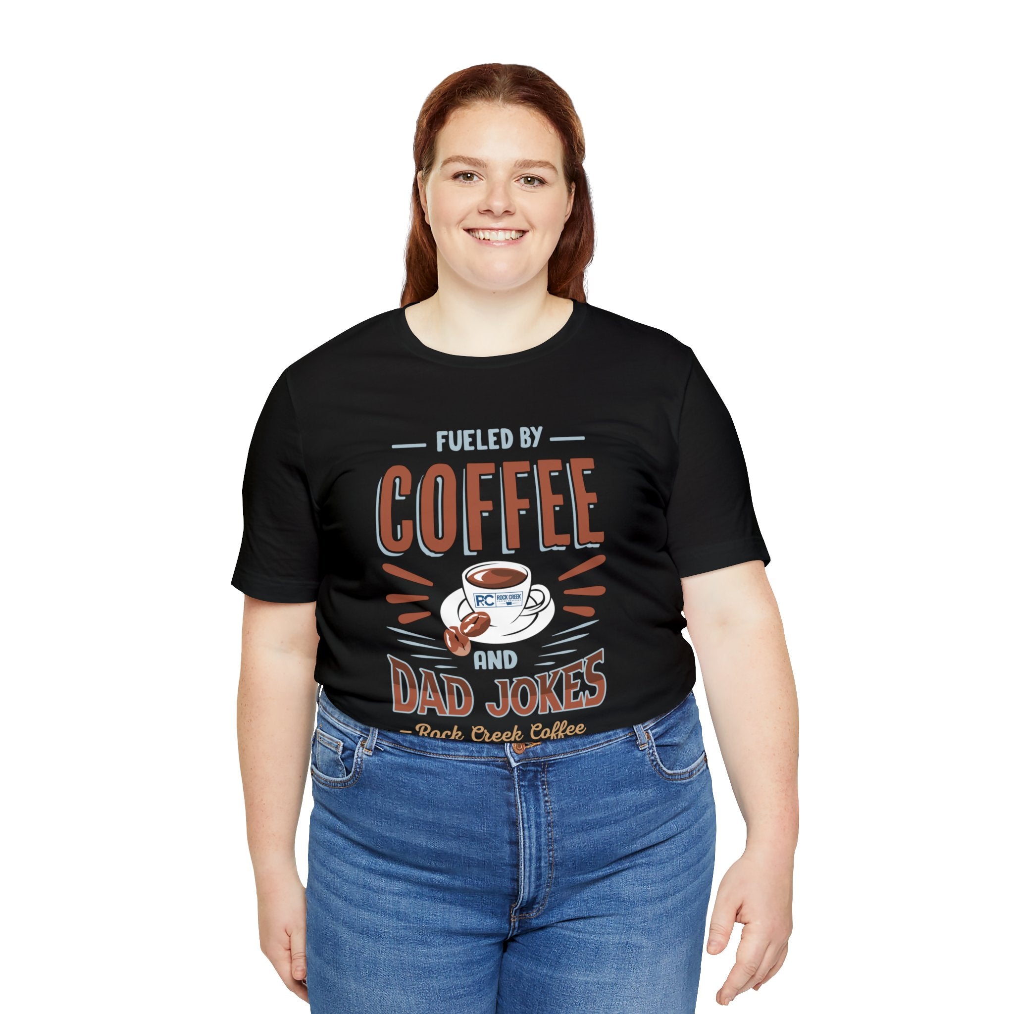 Adult woman smiling while modeling a black short-sleeve T-shirt featuring the “Fueled by Coffee and Dad Jokes” graphic with a coffee cup illustration and Rock Creek Coffee text, shown against a plain white background.