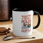White ceramic coffee mug with black handle and black interior featuring the Roasted 818 Miles East of Seattle Rock Creek Coffee design, styled on a wooden surface with a spoon, coffee beans, and a chocolate macaron.