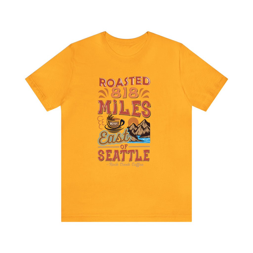Golden yellow short-sleeve T-shirt featuring a colorful “Roasted 818 Miles East of Seattle” graphic with a steaming coffee cup and mountain lake illustration, displayed against a plain white background.