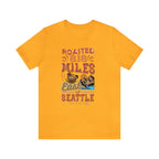Golden yellow short-sleeve T-shirt featuring a colorful “Roasted 818 Miles East of Seattle” graphic with a steaming coffee cup and mountain lake illustration, displayed against a plain white background.