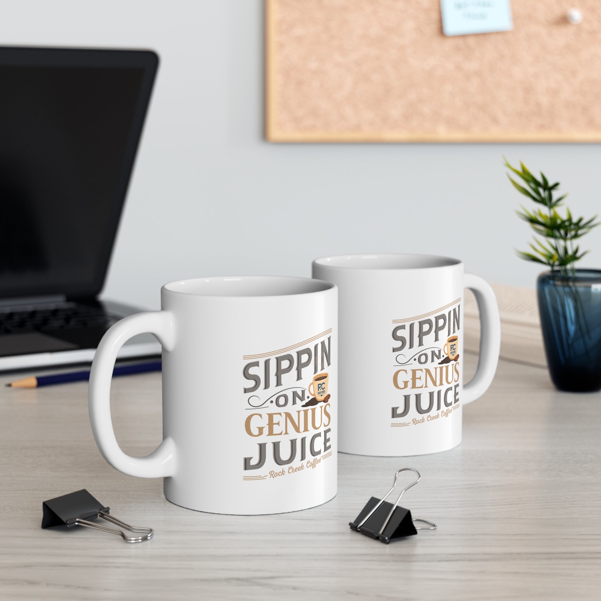 Two white Rock Creek Coffee mugs featuring the phrase “Sippin on Genius Juice” in stylized gray and gold lettering, displayed on a desk with a laptop, binder clips, books, and a small potted plant visible in the background.