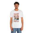Adult man modeling a white short-sleeve T-shirt printed with a colorful graphic reading “Roasted 818 Miles East of Seattle,” featuring a steaming coffee cup and mountain lake illustration, shown against a plain white background.