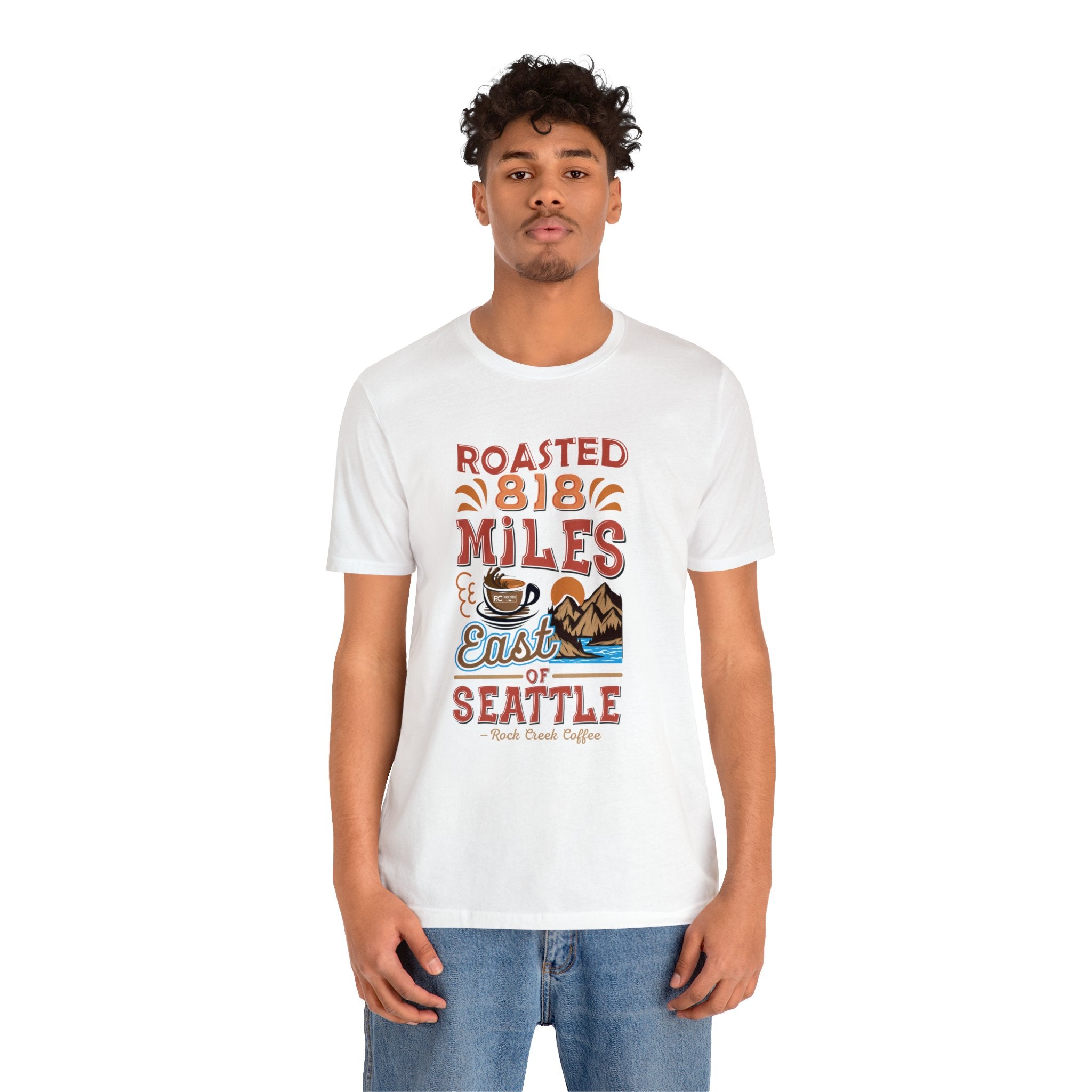 Adult man modeling a white short-sleeve T-shirt printed with a colorful graphic reading “Roasted 818 Miles East of Seattle,” featuring a steaming coffee cup and mountain lake illustration, shown against a plain white background.