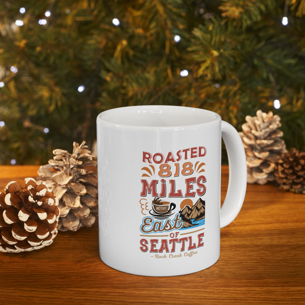 White Rock Creek Coffee mug featuring the colorful “Roasted 818 Miles East of Seattle” design, styled on a wooden surface with pinecones and soft holiday lights in the background.