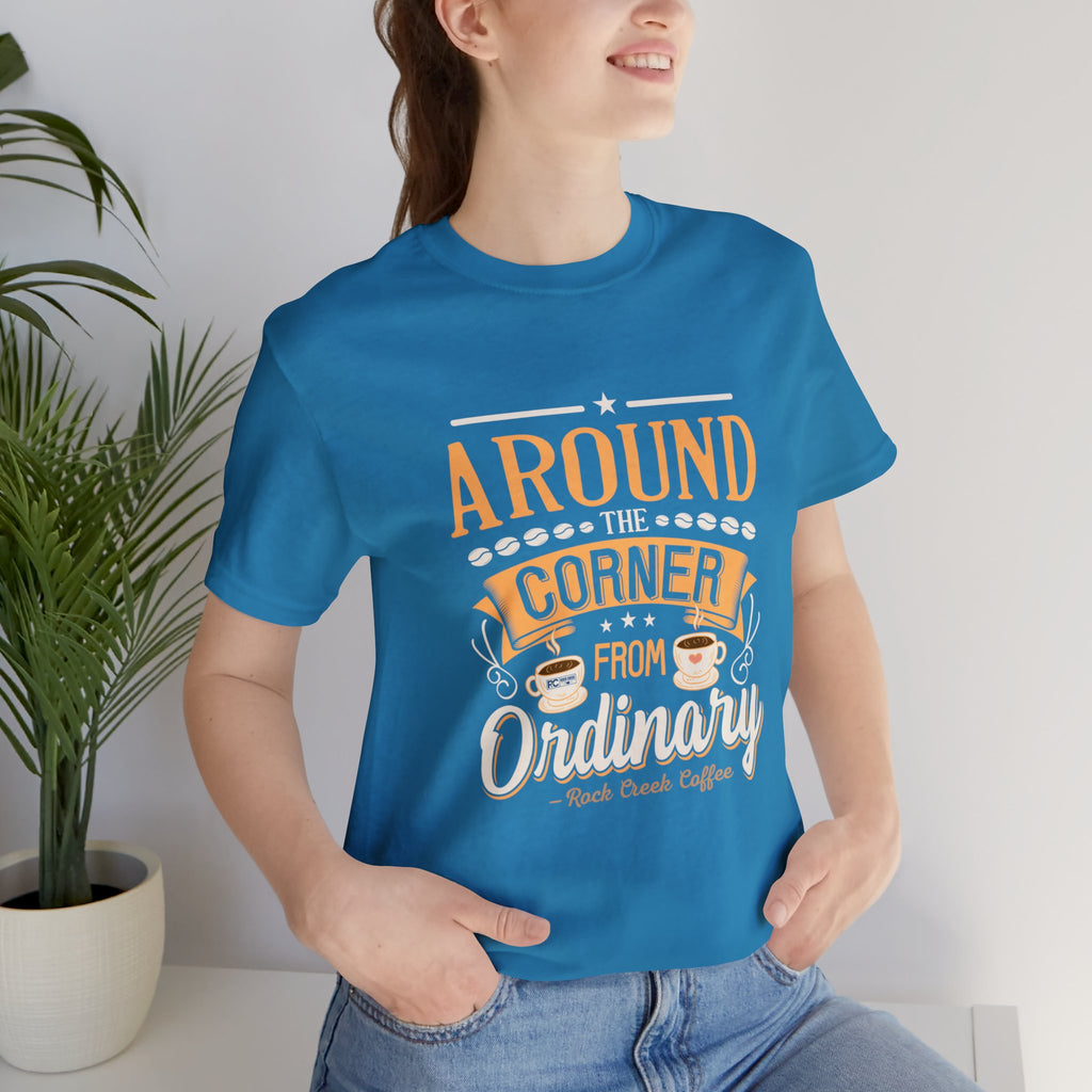 Adult woman modeling an aqua short-sleeve T-shirt featuring the “Around the Corner from Ordinary” graphic in a bold coffee-themed typographic design, photographed indoors beside a potted plant against a light neutral background.