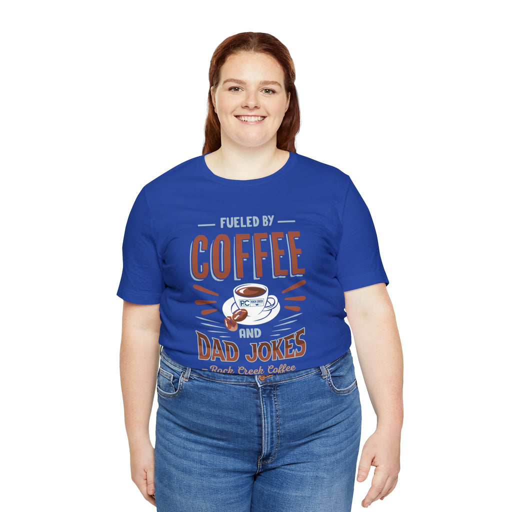 Adult woman smiling while modeling a royal blue short-sleeve T-shirt featuring the “Fueled by Coffee and Dad Jokes” graphic with a coffee cup illustration and Rock Creek Coffee text, shown against a plain white background.