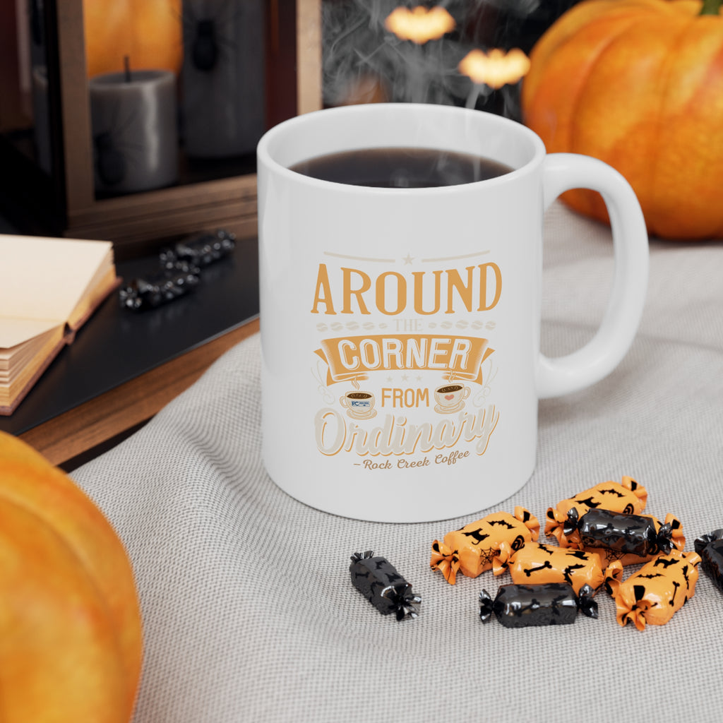 White Rock Creek Coffee mug with “Around the Corner from Ordinary” design in gold and cream lettering, filled with hot coffee and styled with pumpkins, wrapped Halloween candies, candles, and warm seasonal decor.