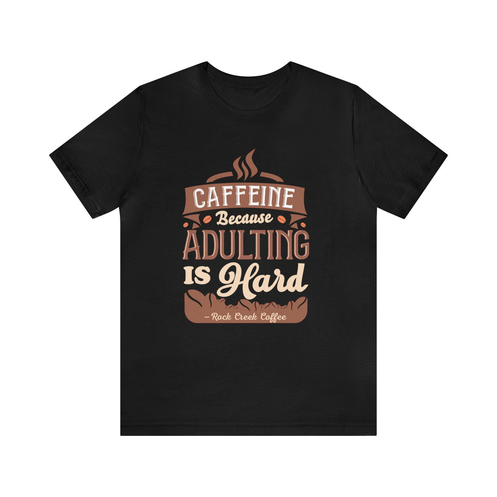 Black short-sleeve T-shirt displayed flat against a plain white background, featuring the phrase “Caffeine Because Adulting Is Hard” in a stacked typographic design with Rock Creek Coffee branding below.