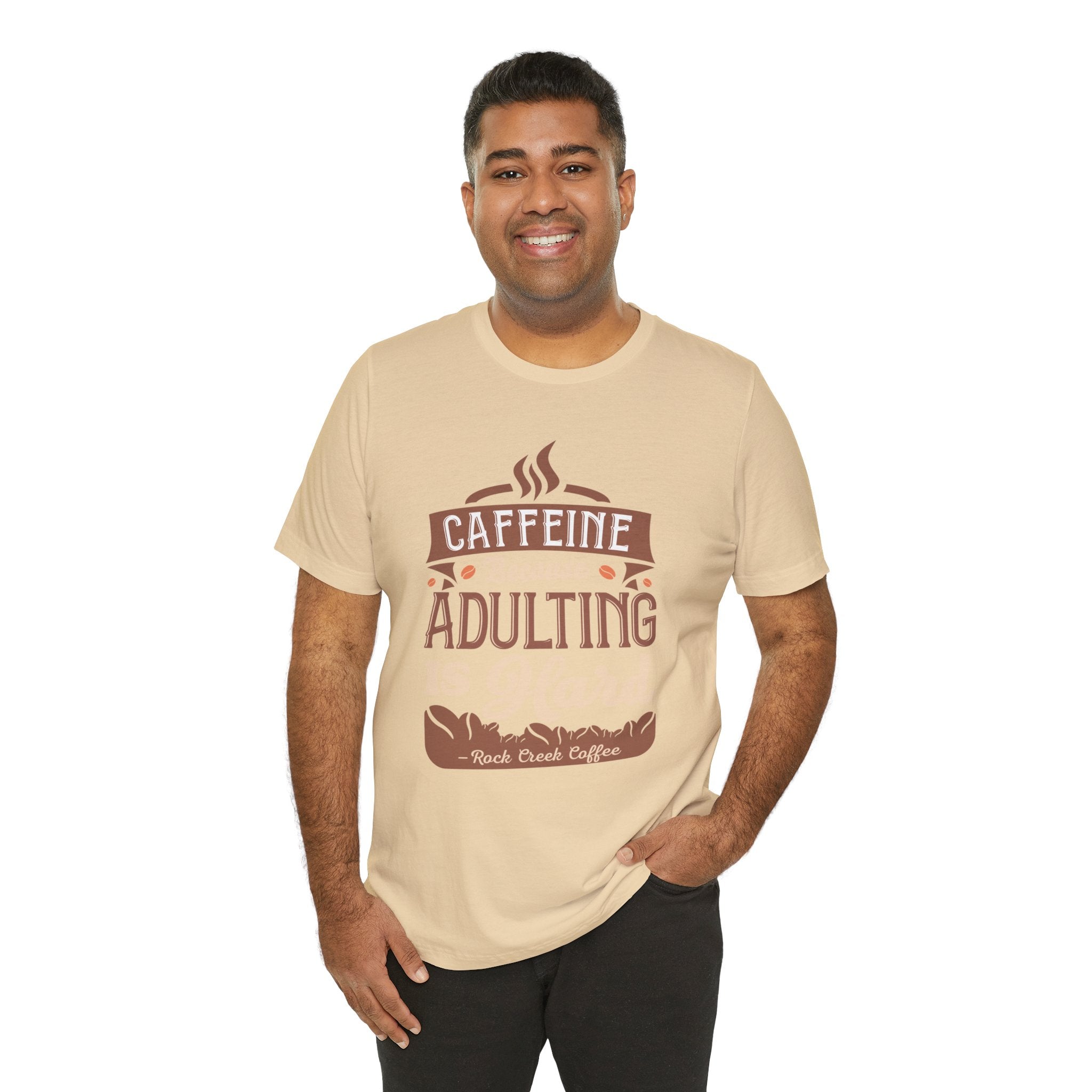 Adult man smiling while modeling a soft cream short-sleeve T-shirt featuring the phrase “Caffeine Because Adulting Is Hard” in a stacked typographic design, shown against a plain white background.