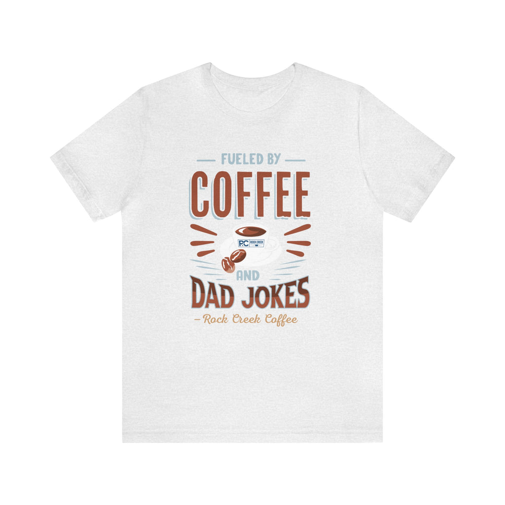 Ash short-sleeve T-shirt displayed flat against a plain white background, featuring the “Fueled by Coffee and Dad Jokes” graphic with a coffee cup illustration and Rock Creek Coffee text below.