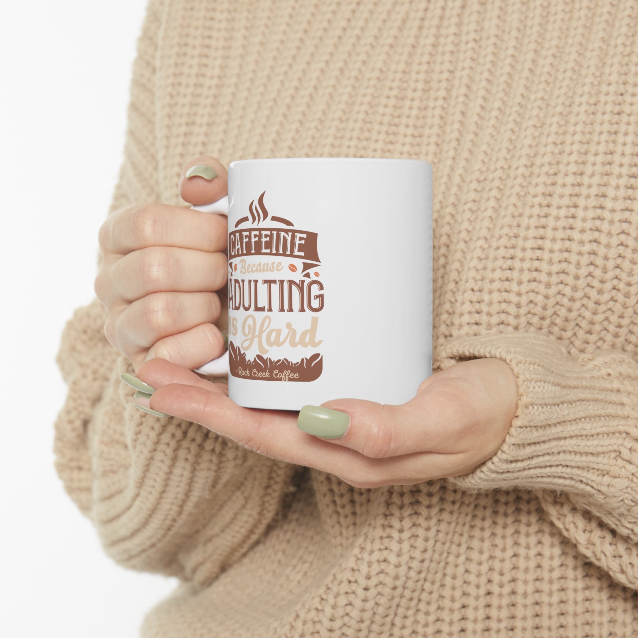 White Rock Creek Coffee mug featuring the phrase “Caffeine Because Adulting Is Hard” in brown and tan lettering, held by a person wearing a beige knit sweater with green painted nails against a plain white background.