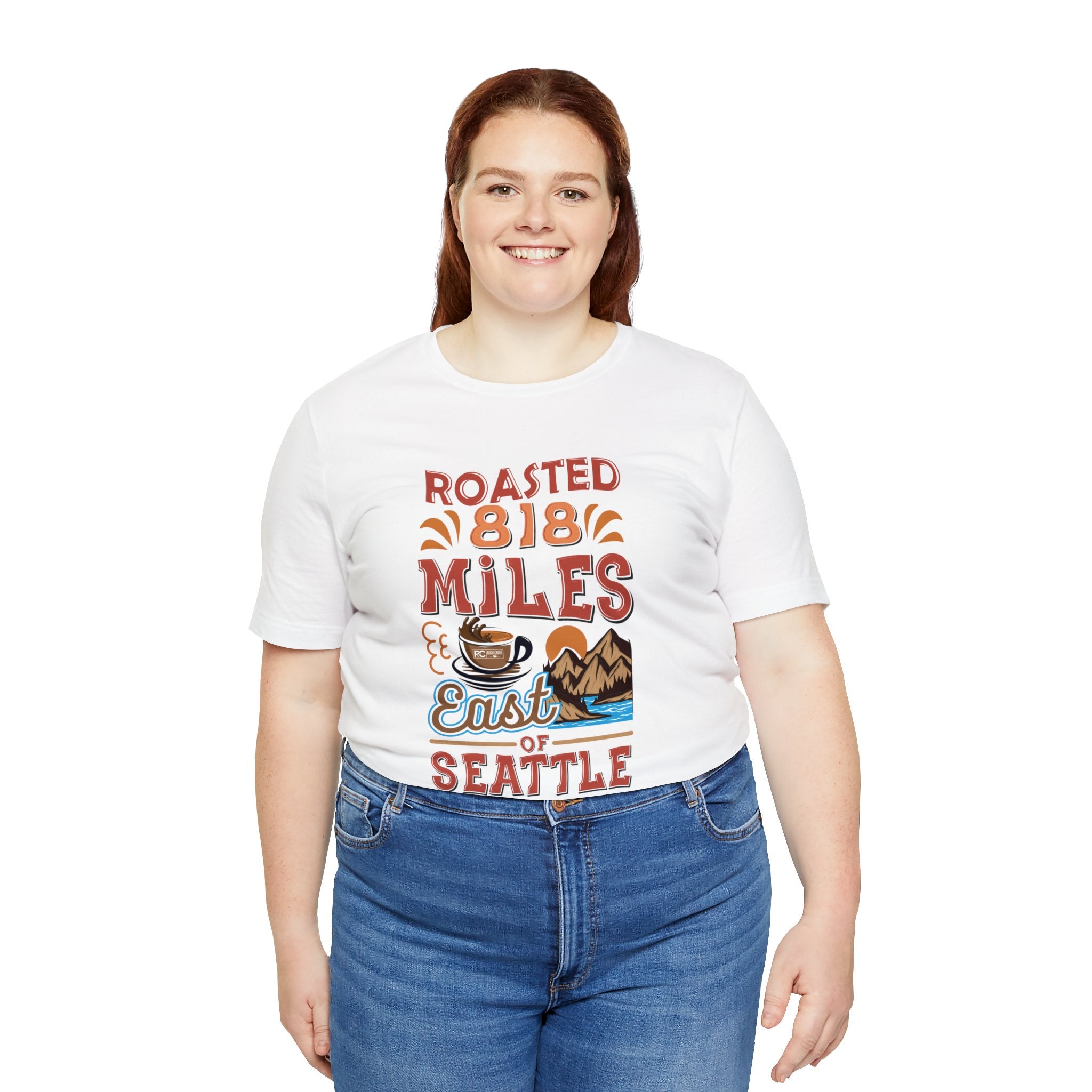 Smiling woman modeling a white short-sleeve T-shirt printed with a colorful coffee-themed graphic that reads “Roasted 818 Miles East of Seattle,” featuring a steaming coffee cup and mountain lake illustration, shown against a plain white background.