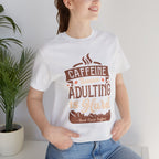 Adult woman modeling an ash short-sleeve T-shirt featuring the “Caffeine Because Adulting Is Hard” graphic in a stacked typographic design, photographed indoors beside a potted plant against a light neutral background.