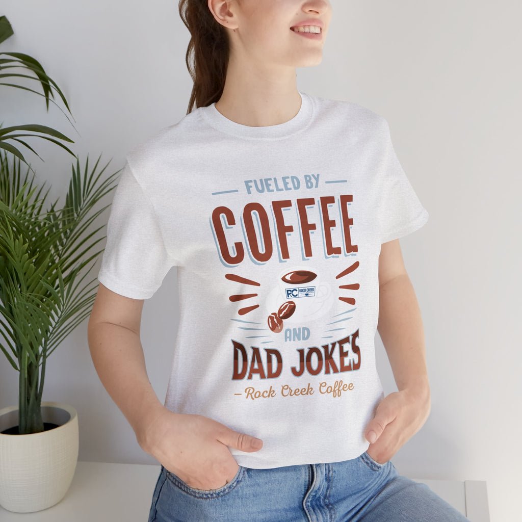Adult woman modeling an ash short-sleeve T-shirt featuring the “Fueled by Coffee and Dad Jokes” graphic with a coffee cup illustration and Rock Creek Coffee text, photographed indoors beside a potted plant against a light neutral background.