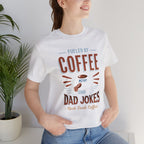 Adult woman modeling an ash short-sleeve T-shirt featuring the “Fueled by Coffee and Dad Jokes” graphic with a coffee cup illustration and Rock Creek Coffee text, photographed indoors beside a potted plant against a light neutral background.