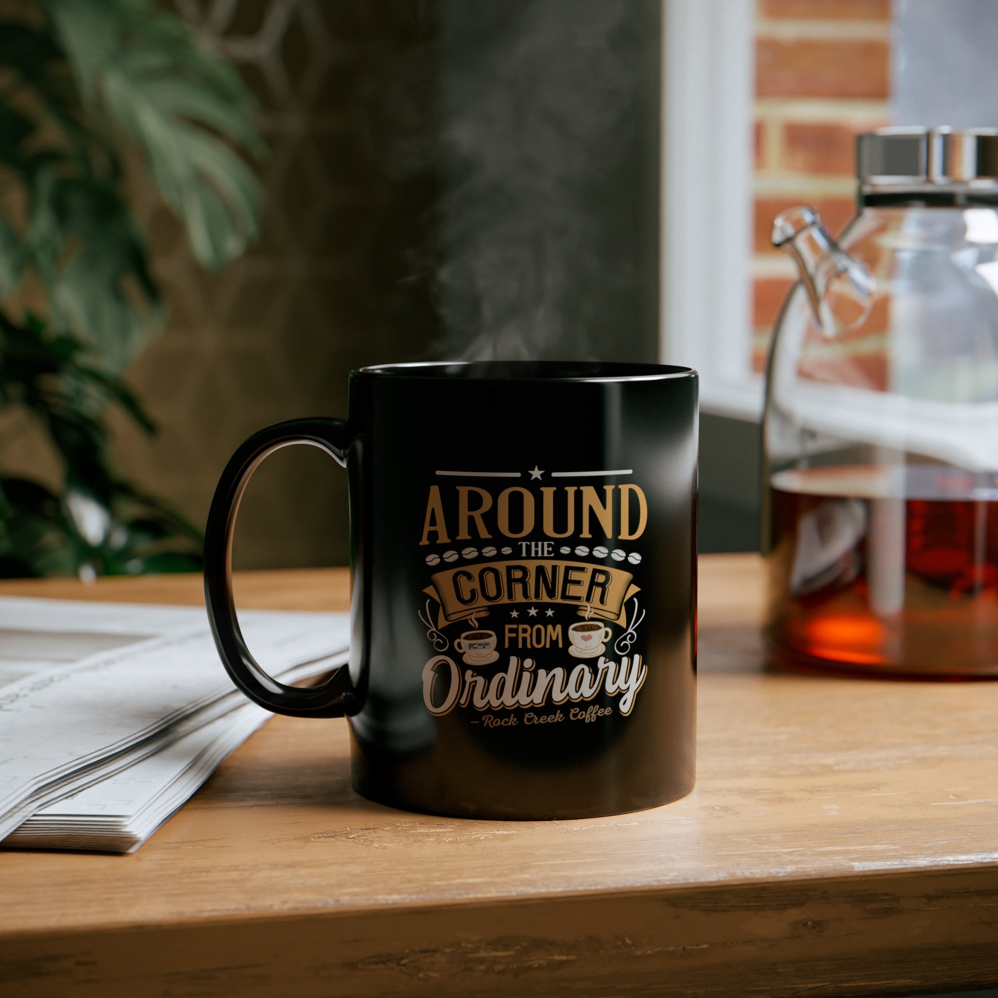 Black ceramic coffee mug featuring the Around the Corner from Ordinary Rock Creek Coffee design in gold and white lettering, filled with hot coffee and steaming, placed on a wooden desk beside a stack of papers with a glass coffee carafe and indoor plant in the background.