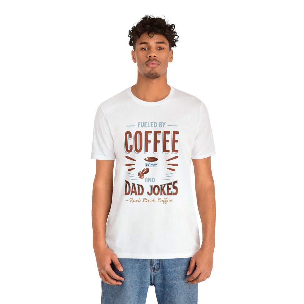 Adult man modeling a white short-sleeve T-shirt featuring the “Fueled by Coffee and Dad Jokes” graphic with a coffee cup illustration and Rock Creek Coffee text, shown against a plain white background.