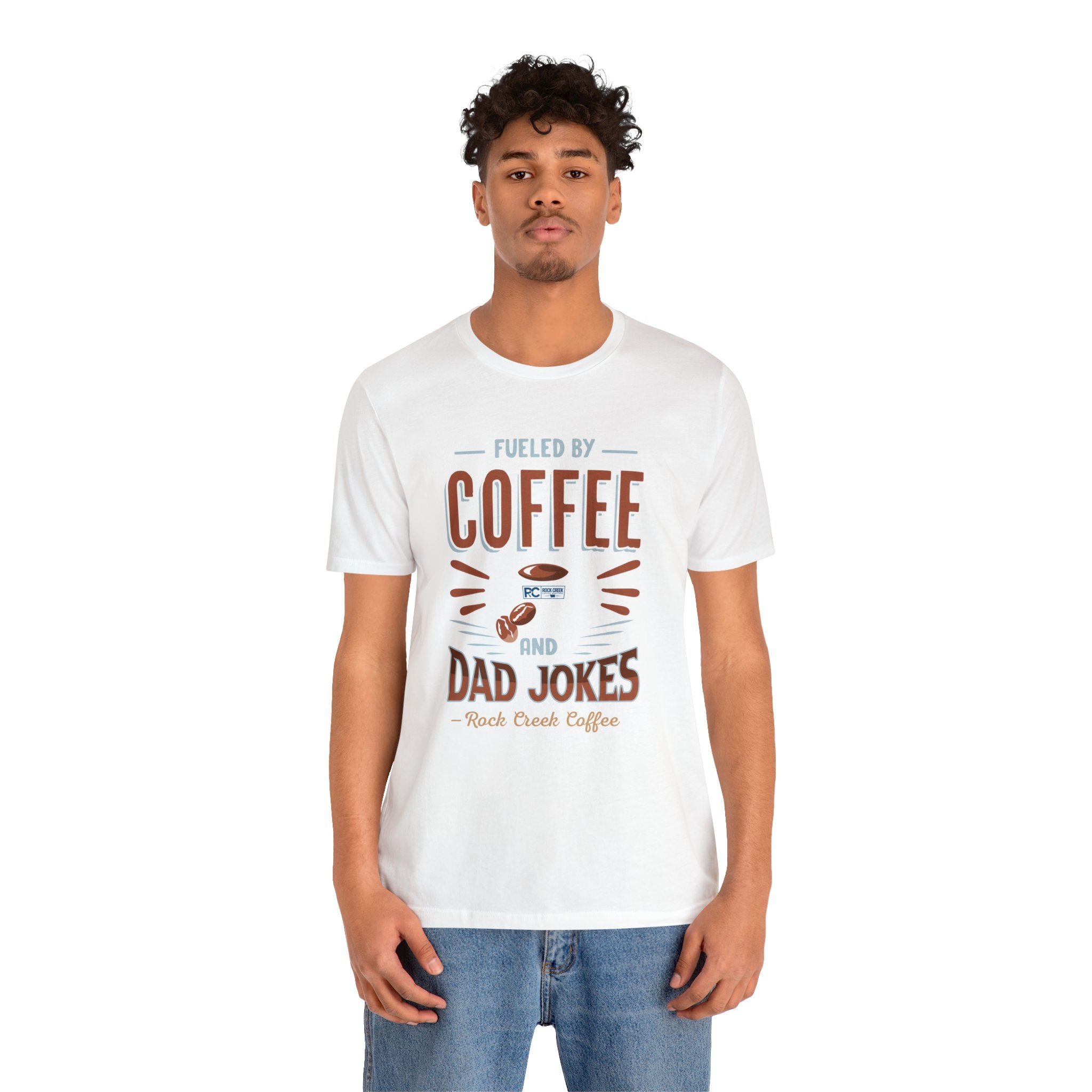 Adult man modeling a white short-sleeve T-shirt featuring the “Fueled by Coffee and Dad Jokes” graphic with a coffee cup illustration and Rock Creek Coffee text, shown against a plain white background.
