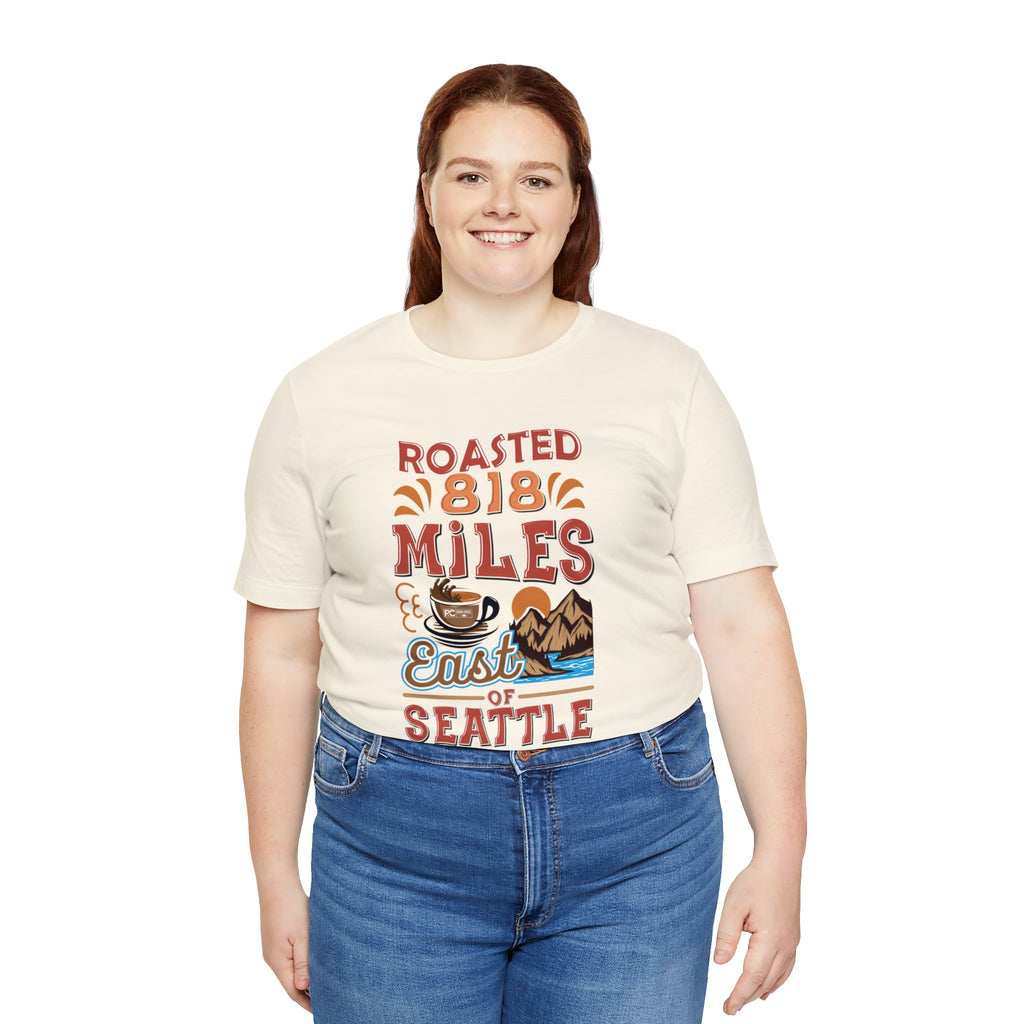 Adult woman smiling while modeling a cream short-sleeve T-shirt featuring the “Roasted 818 Miles East of Seattle” graphic with a steaming coffee cup and mountain lake illustration, shown against a plain white background.