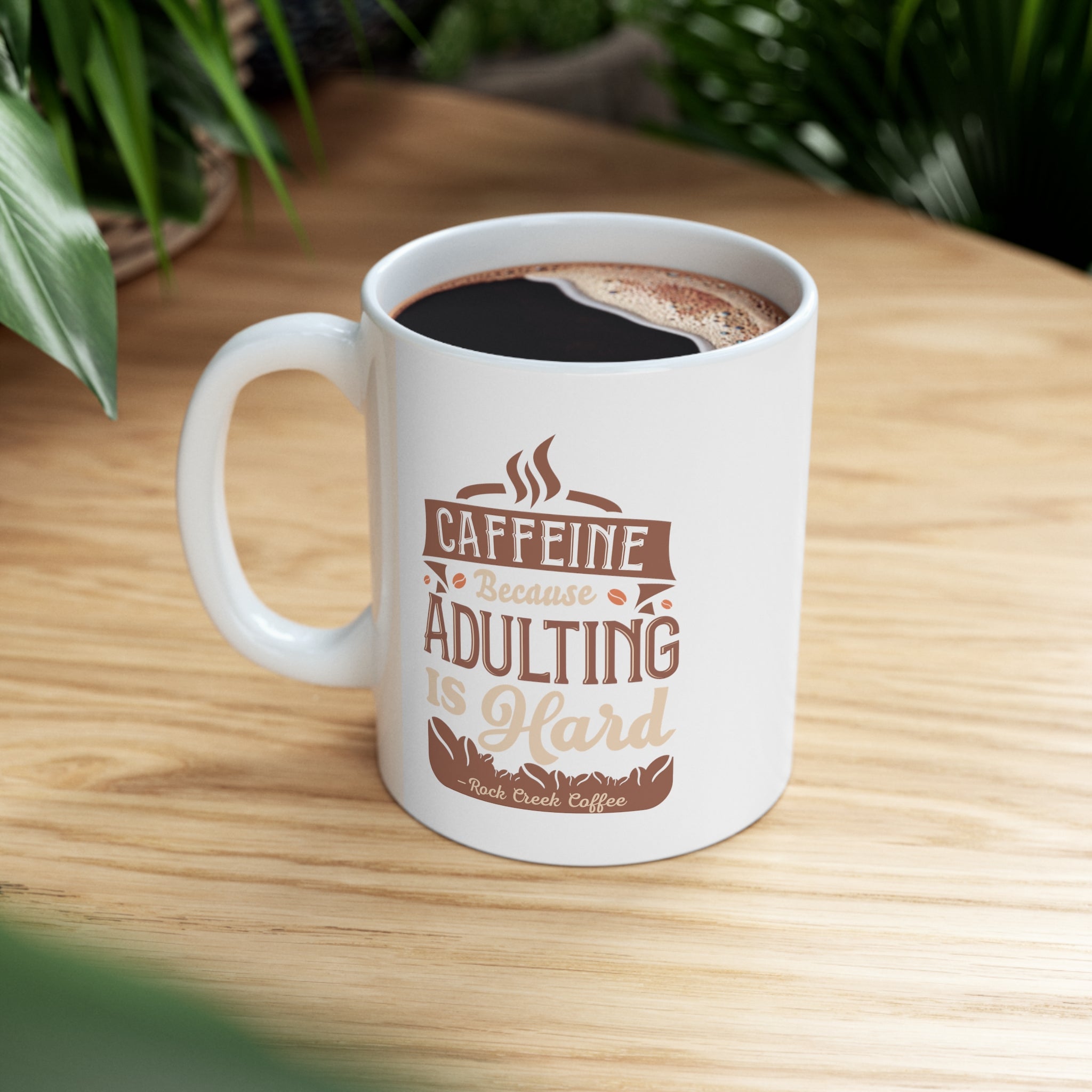 White Rock Creek Coffee mug featuring the phrase “Caffeine Because Adulting Is Hard” in brown and tan lettering, filled with coffee and placed on a wooden table with green plants blurred in the background.