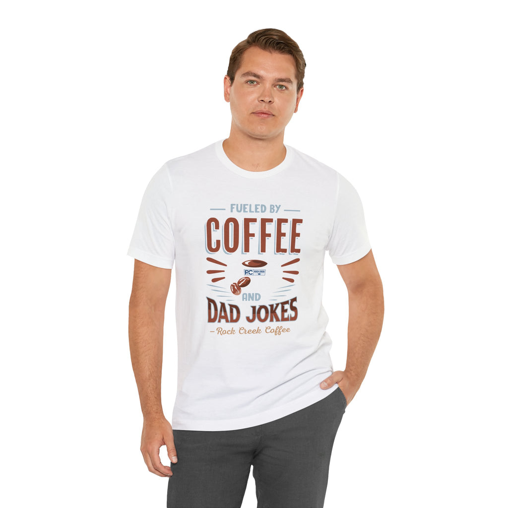 Adult man posing with one hand in his pocket while modeling a white short-sleeve T-shirt featuring the “Fueled by Coffee and Dad Jokes” graphic with a coffee cup illustration and Rock Creek Coffee text, shown against a plain white background.
