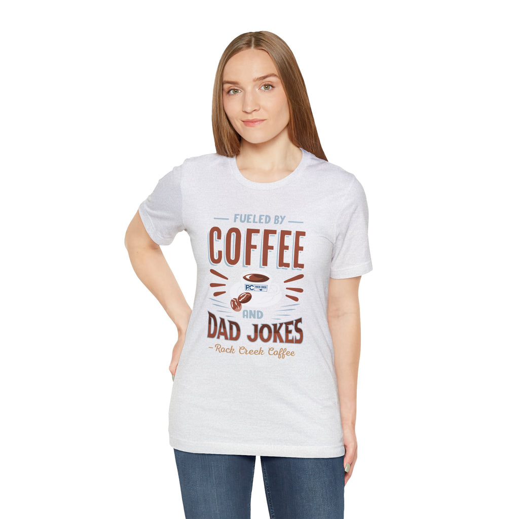 Adult woman modeling an ash short-sleeve T-shirt featuring the “Fueled by Coffee and Dad Jokes” graphic with a coffee cup illustration and Rock Creek Coffee text, shown against a plain white background.