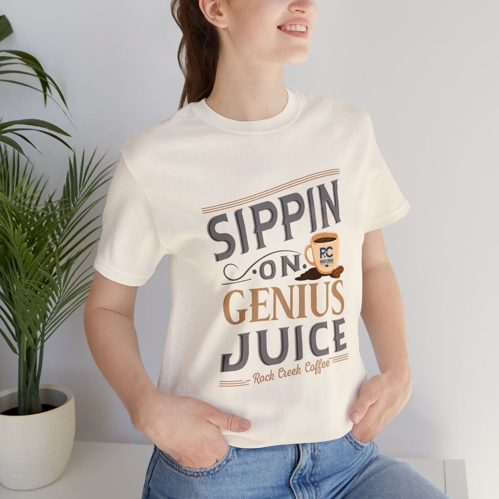 Lifestyle image of an adult woman wearing a cream short-sleeve T-shirt with the “Sippin on Genius Juice” graphic, featuring bold coffee-inspired lettering and a small cup illustration, photographed indoors beside a potted plant.