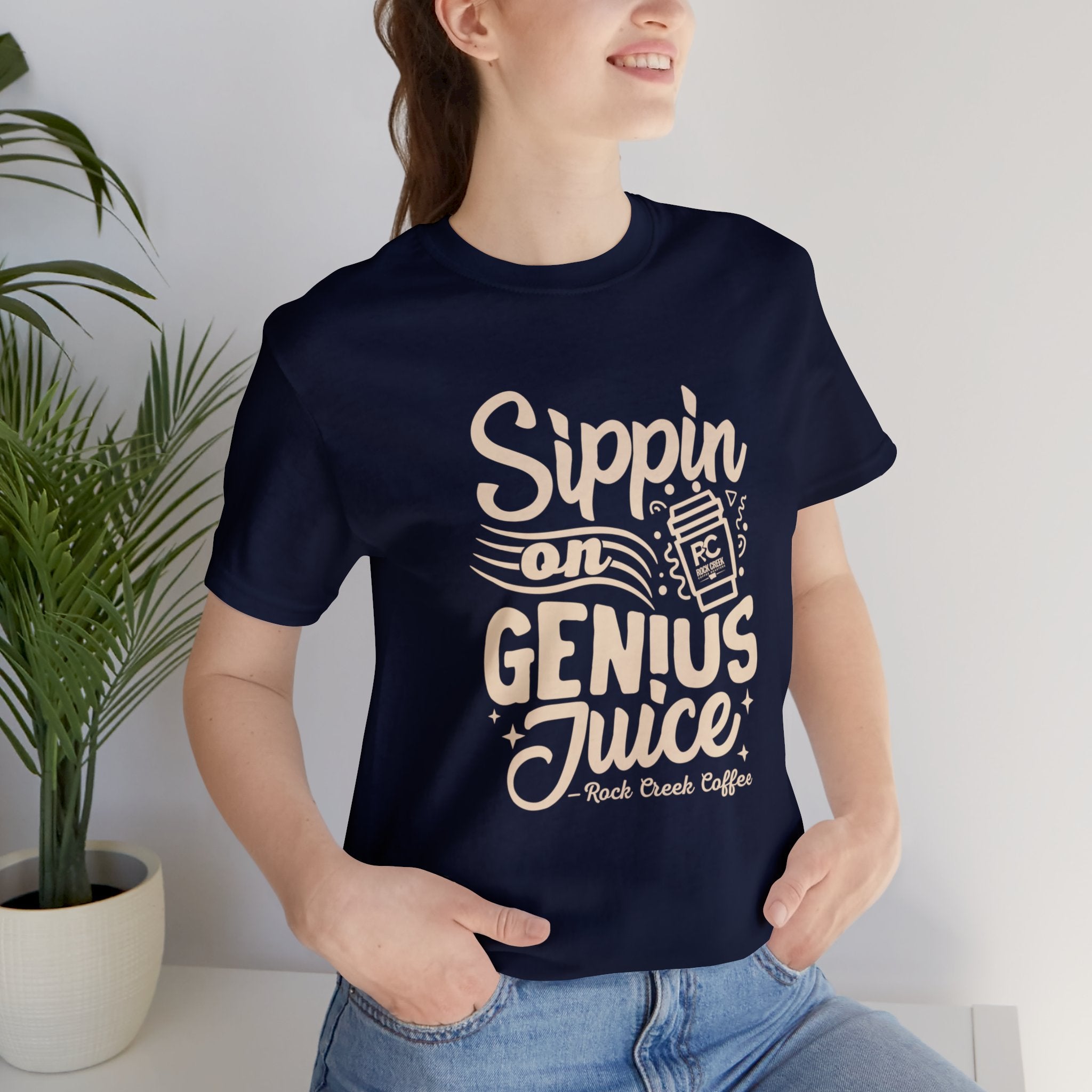 Smiling woman wearing a navy short-sleeve T-shirt printed with the slogan “Sippin on Genius Juice” and a stylized coffee to-go cup graphic, seated casually with a plant visible in the background.