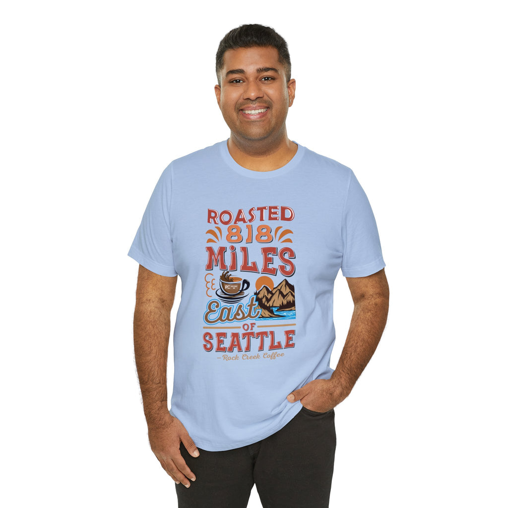 Smiling man modeling a light blue short-sleeve T-shirt printed with a coffee-themed graphic reading “Roasted 818 Miles East of Seattle,” featuring a steaming coffee cup and mountain lake illustration, shown against a plain white background.