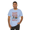 Smiling man modeling a light blue short-sleeve T-shirt printed with a coffee-themed graphic reading “Roasted 818 Miles East of Seattle,” featuring a steaming coffee cup and mountain lake illustration, shown against a plain white background.
