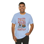 Smiling man modeling a light blue short-sleeve T-shirt printed with a coffee-themed graphic reading “Roasted 818 Miles East of Seattle,” featuring a steaming coffee cup and mountain lake illustration, shown against a plain white background.