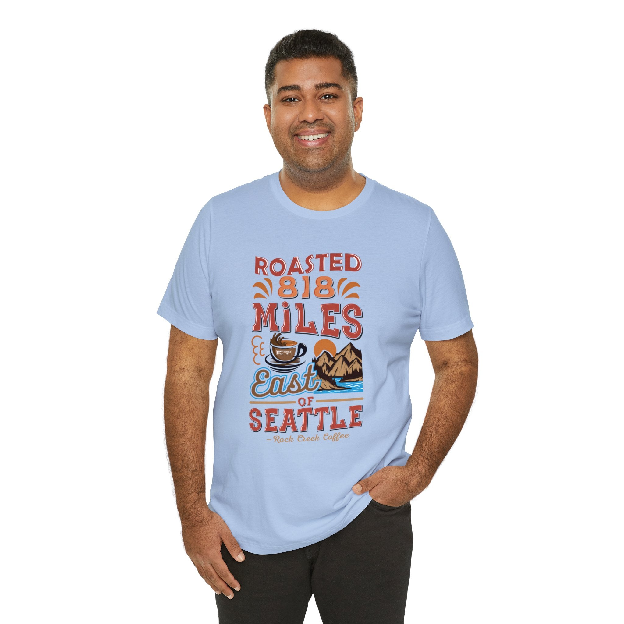 Smiling man modeling a light blue short-sleeve T-shirt printed with a coffee-themed graphic reading “Roasted 818 Miles East of Seattle,” featuring a steaming coffee cup and mountain lake illustration, shown against a plain white background.