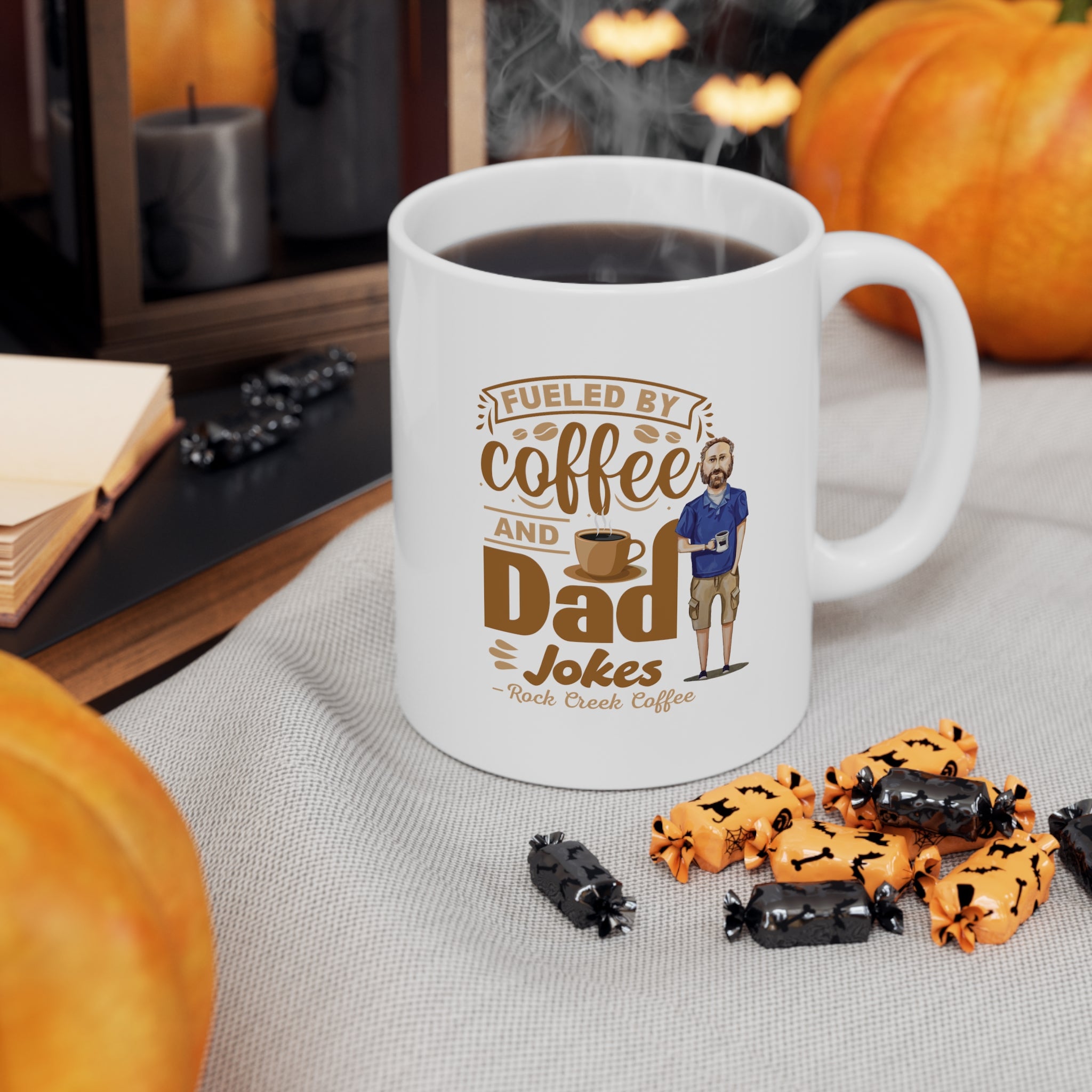 White ceramic coffee mug filled with coffee featuring the text Fueled by Coffee and Dad Jokes Rock Creek Coffee with a cartoon illustration of a man holding a coffee cup, styled with pumpkins, Halloween candy, and cozy fall decor.