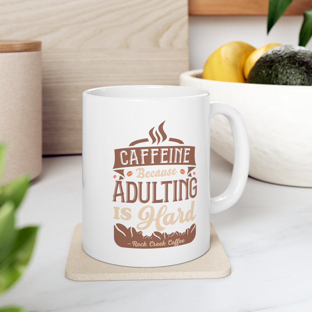 White Rock Creek Coffee mug featuring the phrase “Caffeine Because Adulting Is Hard” in brown and tan lettering, displayed on a kitchen counter with a bowl of lemons and avocados in the background.