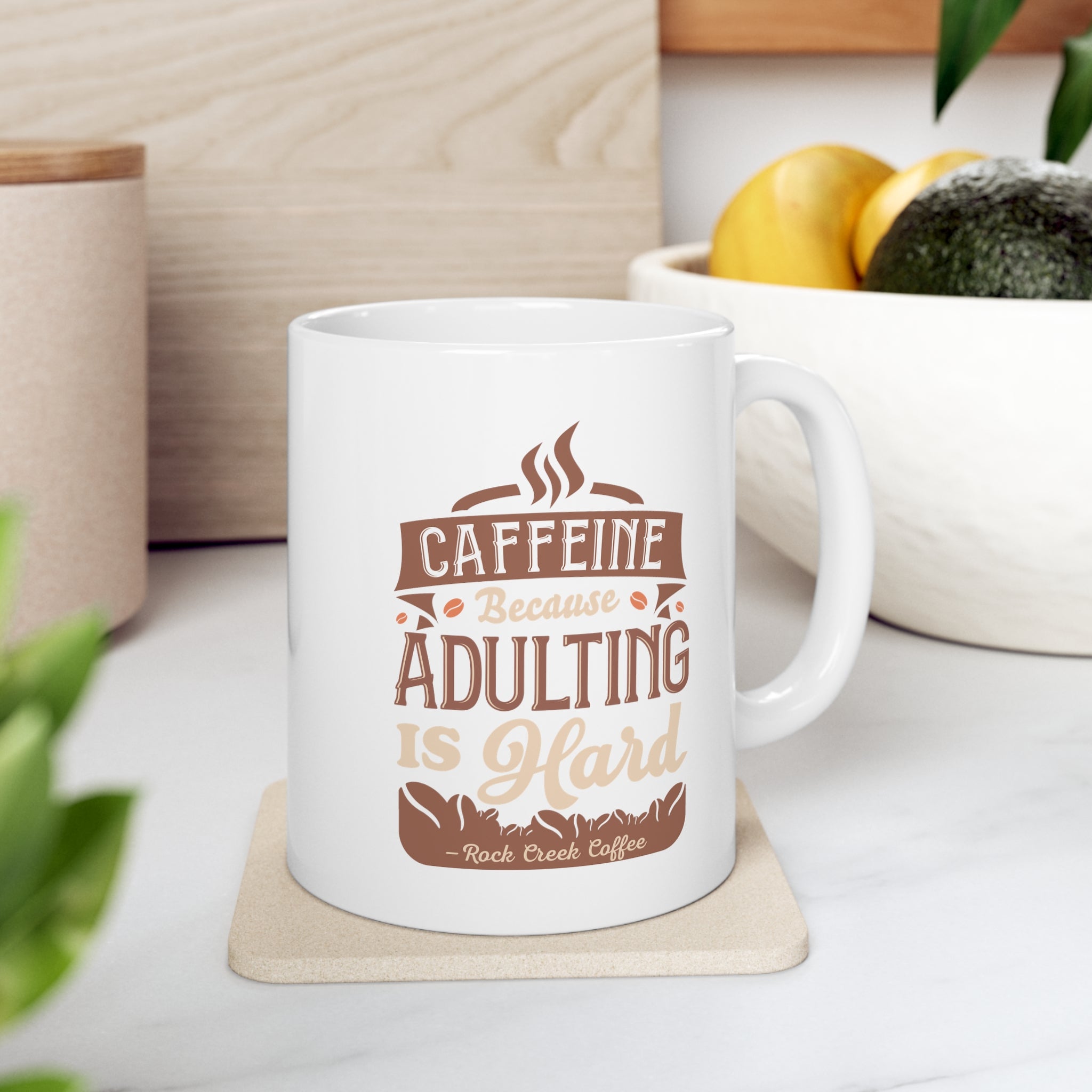 White Rock Creek Coffee mug featuring the phrase “Caffeine Because Adulting Is Hard” in brown and tan lettering, displayed on a kitchen counter with a bowl of lemons and avocados in the background.