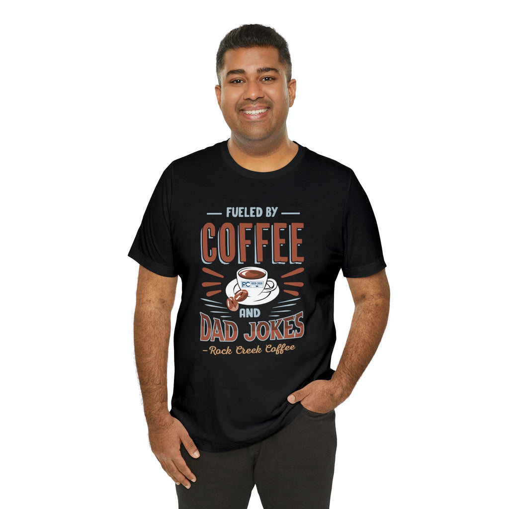 Adult man smiling while modeling a black short-sleeve T-shirt printed with a bold graphic that reads “Fueled by Coffee and Dad Jokes,” featuring coffee-themed artwork, shown against a plain white background.