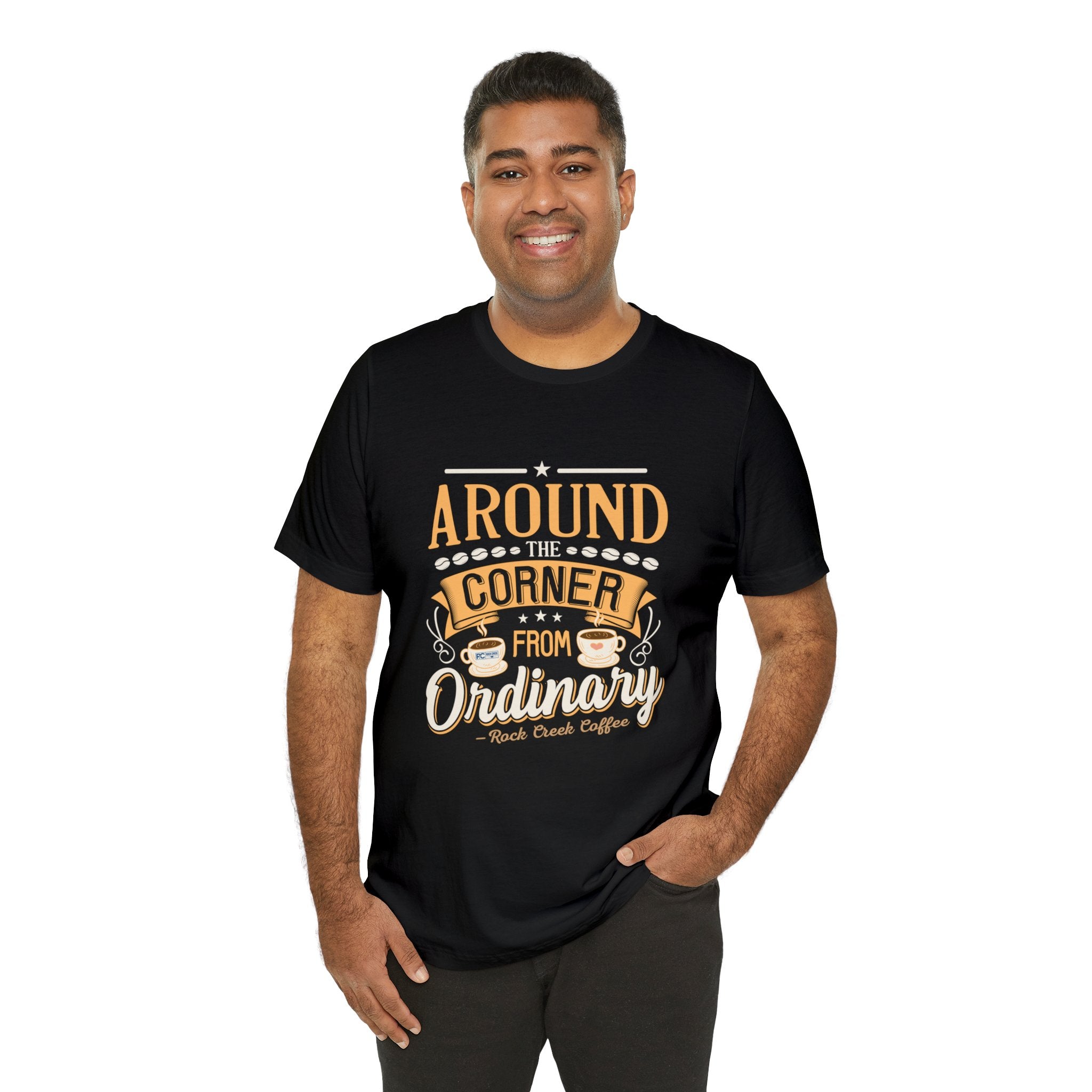 Adult man smiling while modeling a black short-sleeve T-shirt featuring the “Around the Corner from Ordinary” graphic in a bold coffee-themed typographic design, shown against a plain white background.