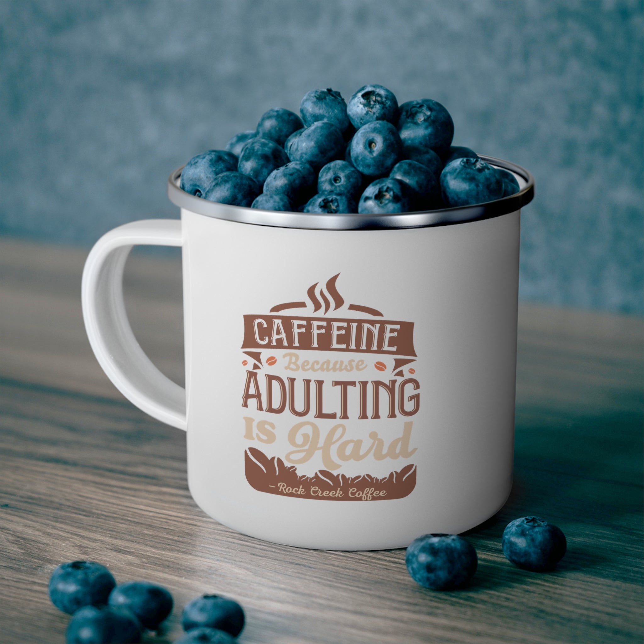 White enamel-style coffee mug with silver rim and handle, printed with “Caffeine Because Adulting Is Hard – Rock Creek Coffee,” filled with fresh blueberries and set on a wooden table with a few berries scattered nearby.