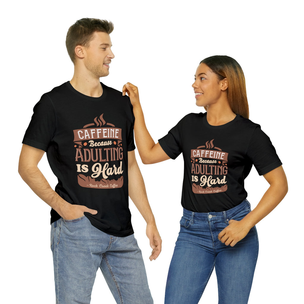 Adult man and woman modeling matching black short-sleeve T-shirts featuring the “Caffeine Because Adulting Is Hard” graphic in a stacked typographic design, posed together against a plain white background.