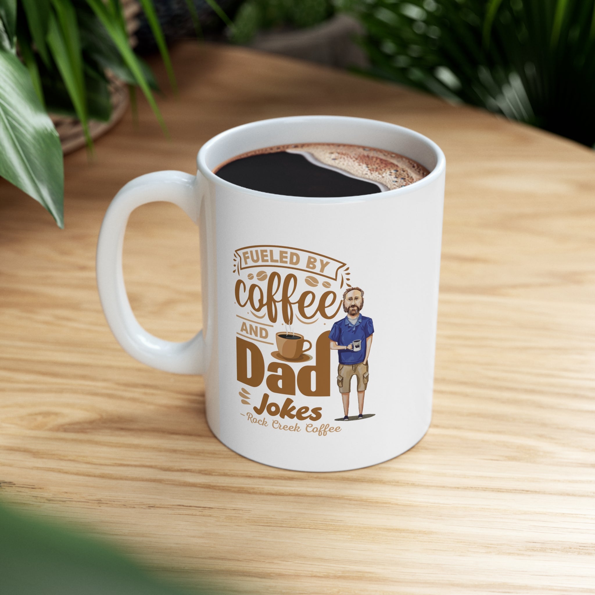 White ceramic coffee mug filled with coffee featuring the text Fueled by Coffee and Dad Jokes Rock Creek Coffee with a cartoon illustration of a man holding a coffee cup, placed on a wooden table with green plants in the background.