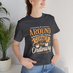 Adult woman modeling an asphalt gray short-sleeve T-shirt featuring the “Around the Corner from Ordinary” graphic in a bold coffee-themed typographic design, photographed indoors beside a potted plant against a light neutral background.