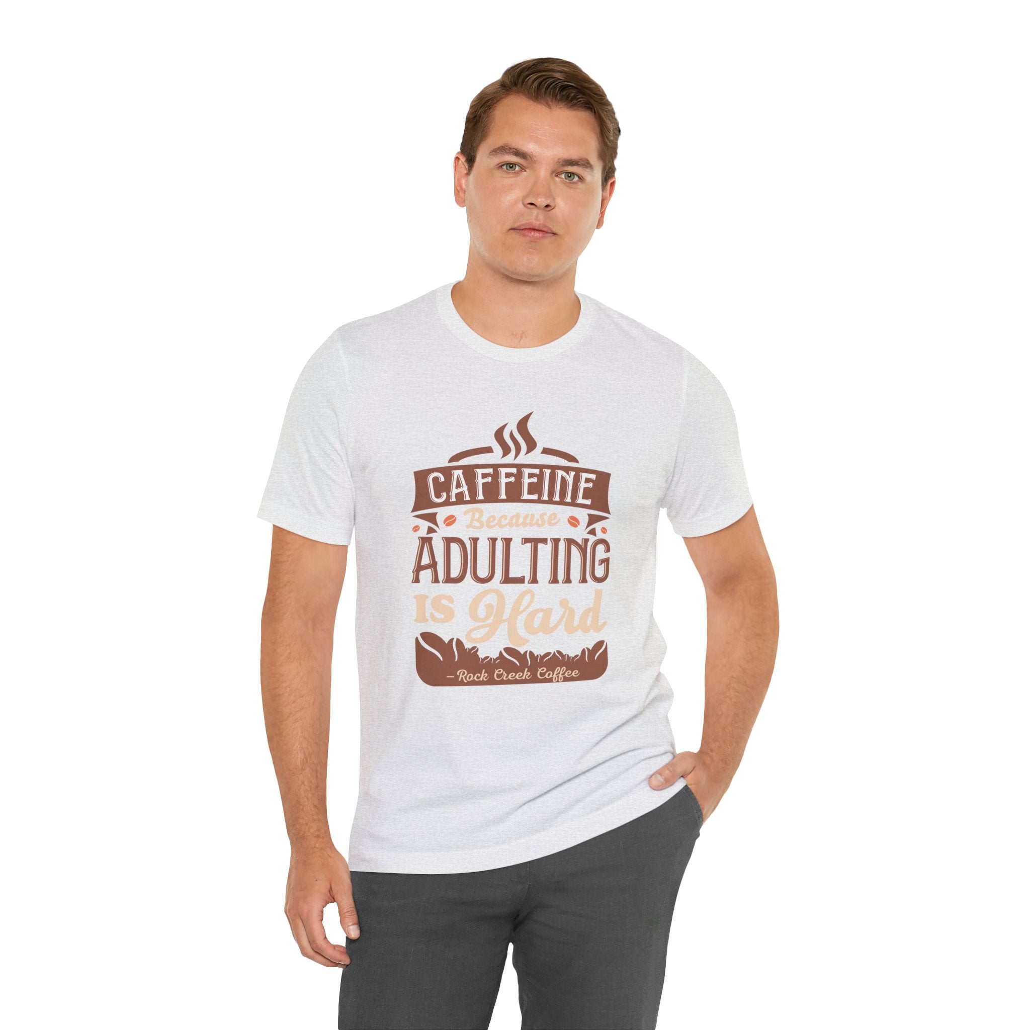 Adult man modeling an ash short-sleeve T-shirt featuring the “Caffeine Because Adulting Is Hard” graphic in a stacked typographic design, shown against a plain white background.