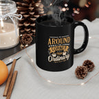 Black ceramic coffee mug with Around the Corner from Ordinary Rock Creek Coffee design in gold and white lettering, filled with hot coffee and steam rising, surrounded by a cozy setting with pinecones, cinnamon sticks, a lit candle in a glass jar, and warm string lights.