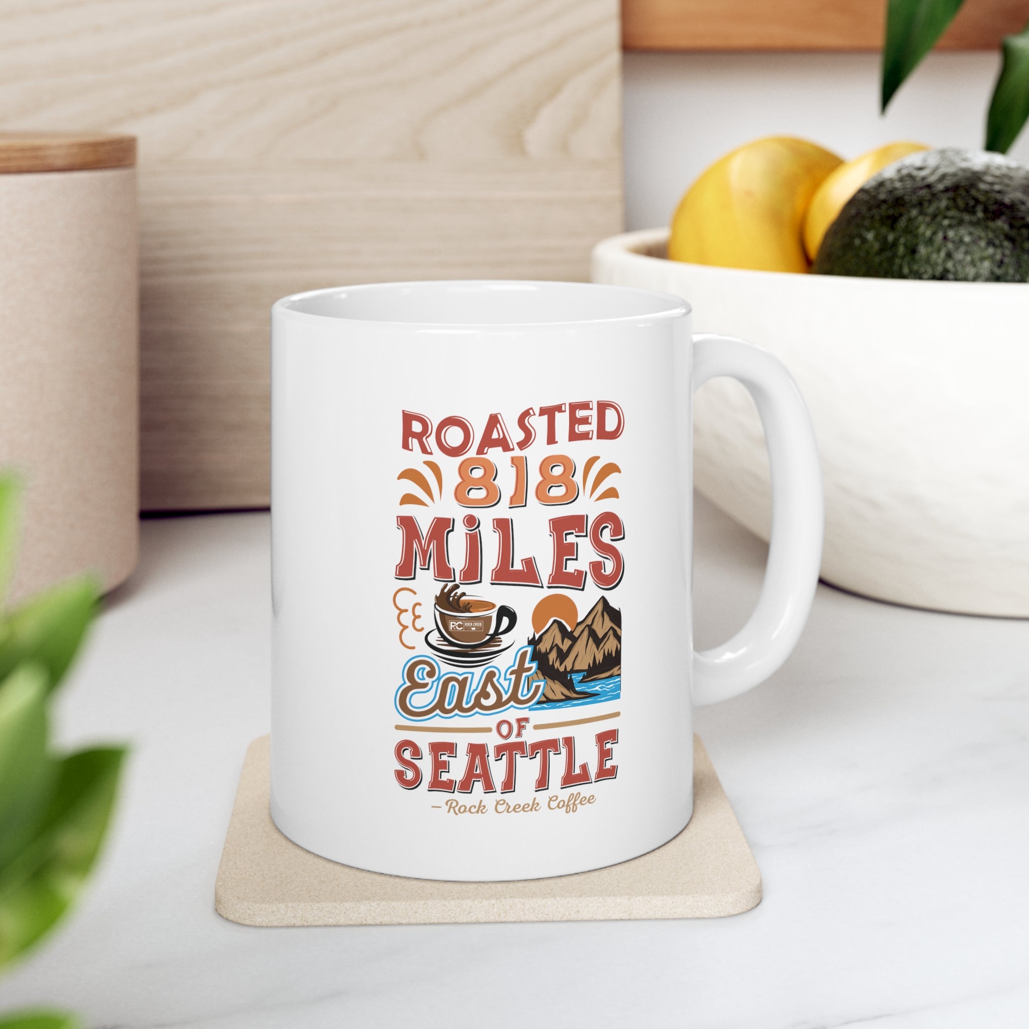 White Rock Creek Coffee mug featuring the colorful “Roasted 818 Miles East of Seattle” design, displayed on a kitchen counter with a bowl of lemons and avocados in the background.