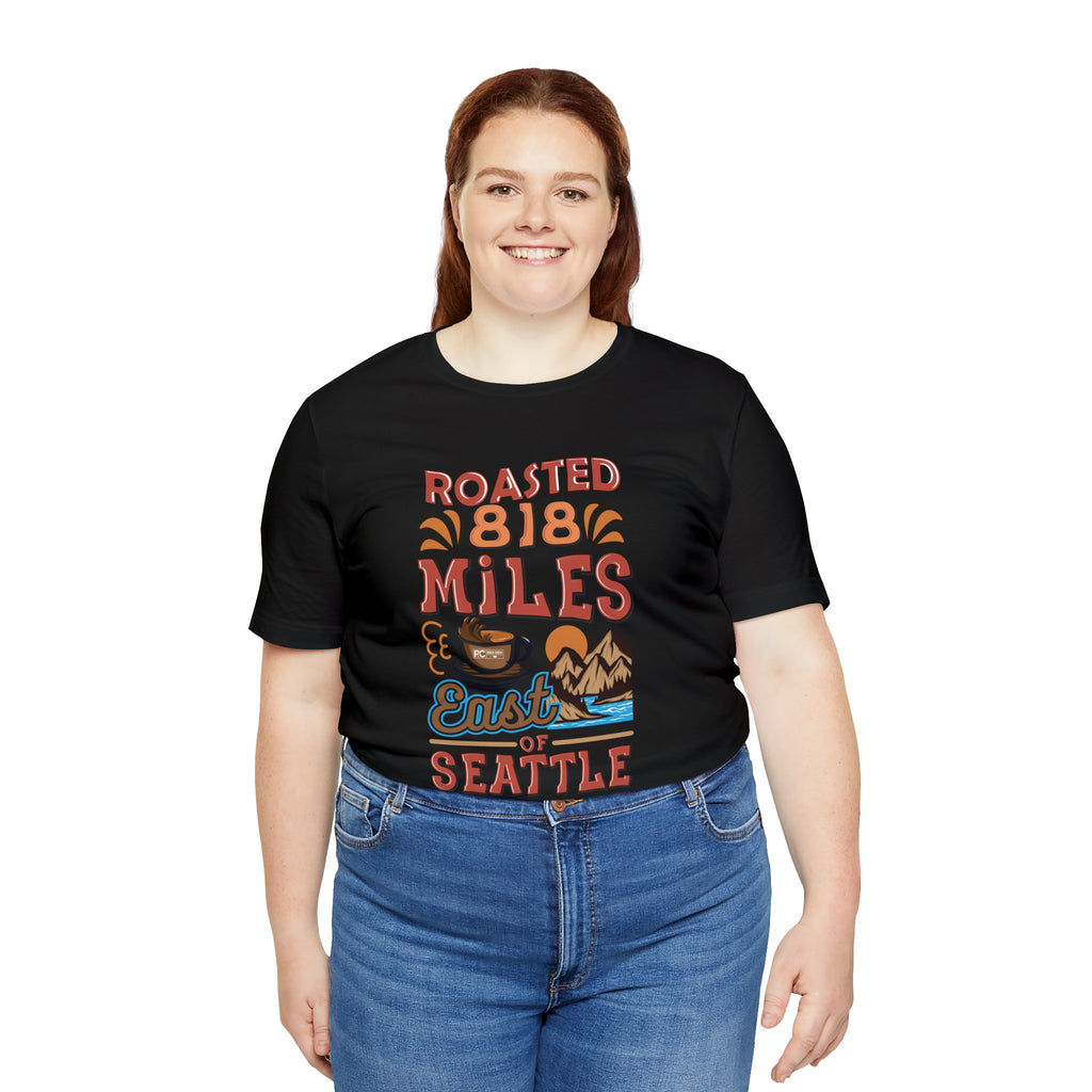 Adult woman modeling a black short-sleeve T-shirt printed with a colorful graphic that reads “Roasted 818 Miles East of Seattle,” featuring a steaming coffee cup and mountain lake illustration, shown against a plain white background.
