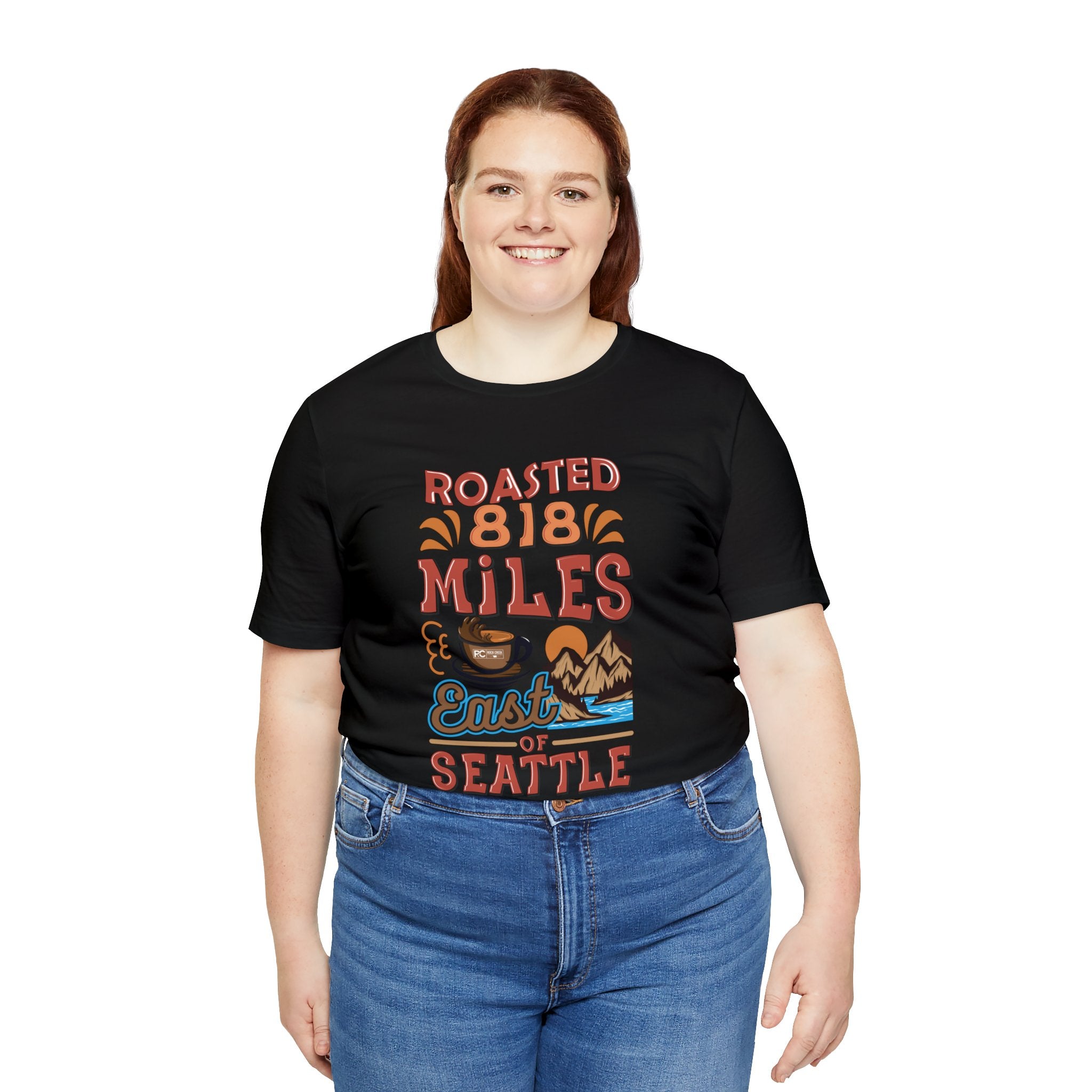 Adult woman modeling a black short-sleeve T-shirt printed with a colorful graphic that reads “Roasted 818 Miles East of Seattle,” featuring a steaming coffee cup and mountain lake illustration, shown against a plain white background.