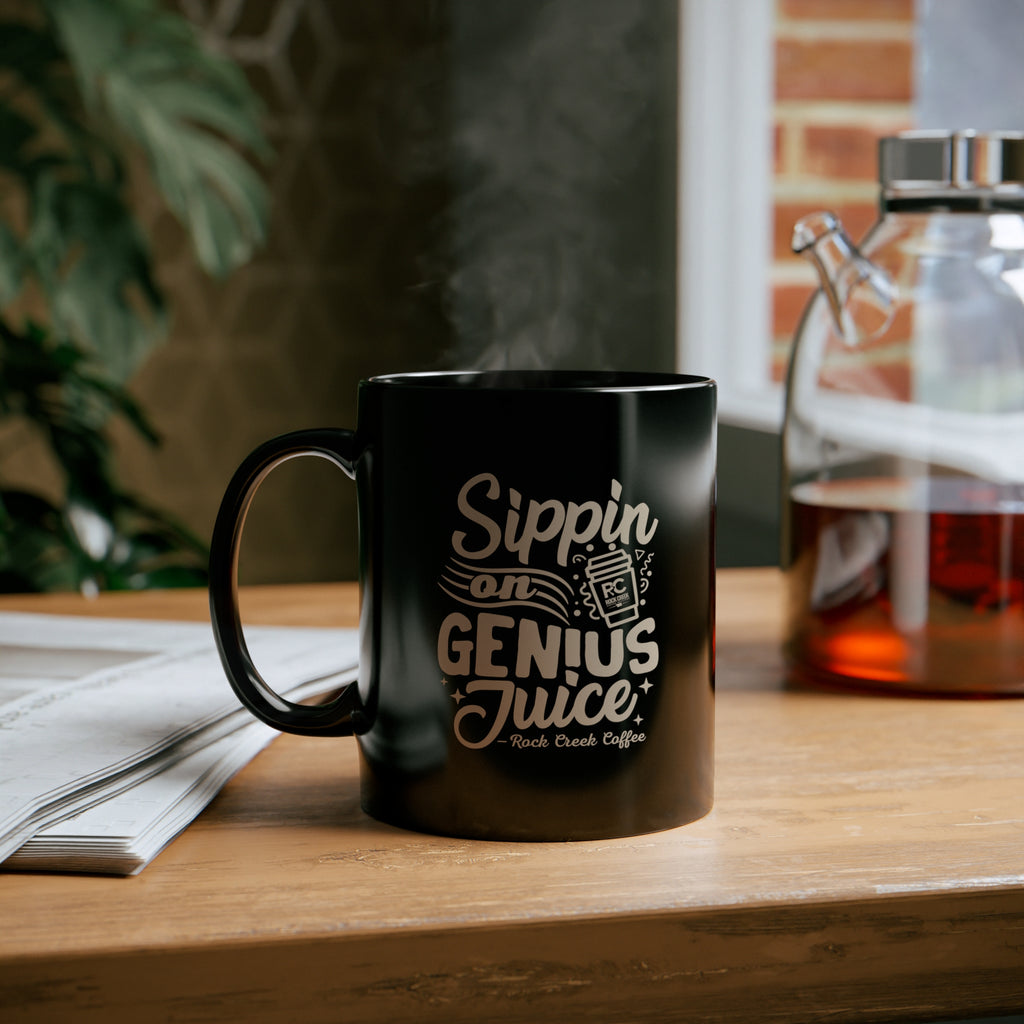 Black Rock Creek Coffee mug with “Sippin on Genius Juice” design in light lettering, filled with steaming coffee and placed on a wooden table beside an open notebook and a glass coffee carafe in a cozy indoor setting.